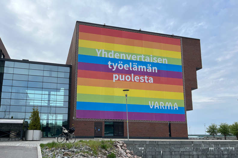 Toimiston ulkoseinällä suuri sateenkaaren värinen seinälakana, jossa teksti "Yhdenvertaisen työelämän puolesta" ja Varman logo, taustalla pilvinen taivas.